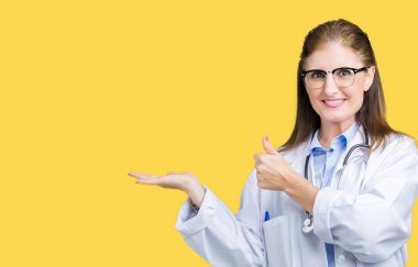 Orta yaş olgun doktor kadın üzerinde tıbbi bir palto giyiyor arka plan gösteren palm el ve iş tamam jestle başparmak yukarıya, mutlu ve neşeli gülümseyen izole
