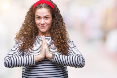 Ellerle birlikte AF emin gülümseyen için soran dua izole arka plan üzerinde kazak giyen güzel esmer kıvırcık saçlı Genç kız çizgili.