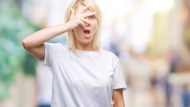 Mahcup ifadeyle parmaklarımın arasından seyir yüz ve gözler el ile kapsayan şok bakmak izole arka plan üzerinde beyaz t-shirt giyen genç güzel sarışın kadın.