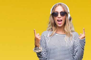 Kulaklık ve güneş gözlüğü izole arka plan üzerinde giyen genç güzel sarışın kadın hayrete ve yukarıya bakıyor ve parmakları ile işaret ve silah kaldırdı şaşırttı.