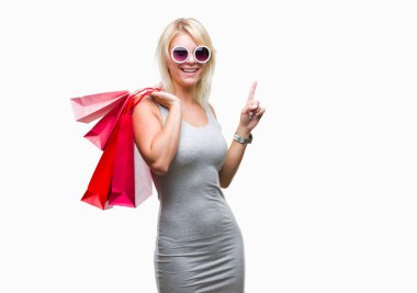 Genç güzel sarışın kadın alışveriş alışveriş torbaları üzerinde satış izole arka plan üzerinde tutan bir fikir ya da soru işaret parmağı gülen yüz, bir numara ile şaşırttı