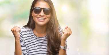 Genç güzel Arap kadın izole arka plan kutlama üzerinde güneş gözlüğü takıyor şaşırttı ve başarı için kaldırdı kollarını açarak hayret ve gözleri açık. Kazanan kavramı.