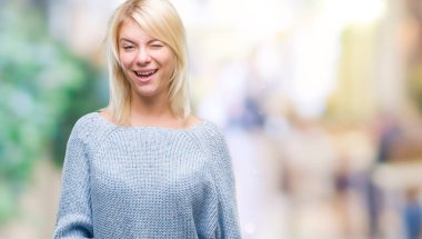 Seksi bir ifadeyle, neşeli ve mutlu yüzünü kameraya bakarak göz kırpıyor izole arka plan üzerinde kış kazak giyen genç güzel sarışın kadın.