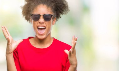 Genç afro Amerikalı kadın deli ve silah ile başarı için deli kutluyor izole arka plan üzerinde güneş gözlüğü takıyor kaldırdı ve heyecanlı çığlık gözler kapalı. Kazanan kavramı