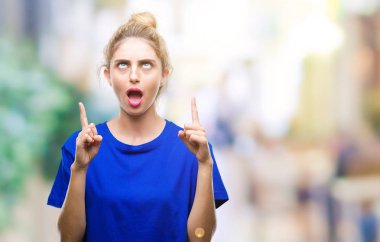 İzole arka plan üzerinde mavi t-shirt giyen genç güzel sarışın ve mavi gözlü kadın hayrete ve yukarıya bakıp parmakları ile işaret şaşırttı ve silah kaldırdı.