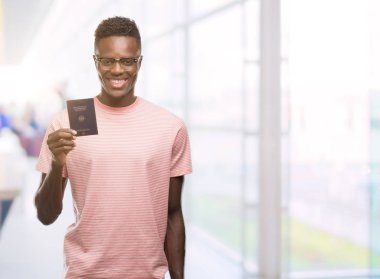 Alman pasaportu ile ayakta ve diş gösterilen kendine güvenen bir gülümsemeyle gülümseyerek mutlu bir yüz tutan genç Afro-Amerikan adam