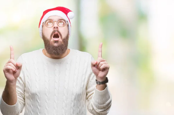 孤立した背景にクリスマスの帽子とメガネを着て若い白人男びっくり驚いて見上げると指で指していると腕を上げた — ストック写真