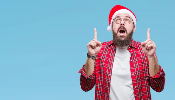 年轻的高加索人在孤立的背景下戴着圣诞帽 惊讶地抬起头来 用手指指指点点 举起双臂 — 图库照片