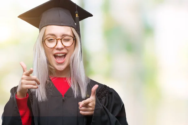 若いブロンドの女性は 幸せと面白い顔でカメラに指を指して孤立の背景に大学院の制服を着ています 良いエネルギーとヴァイブス — ストック写真