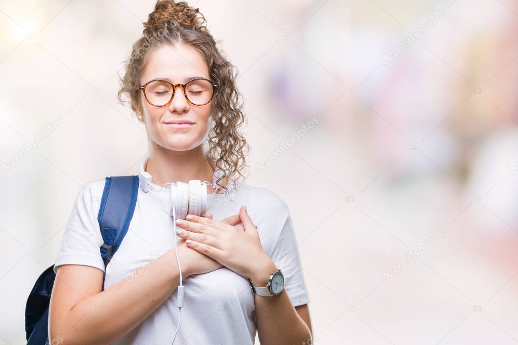 Young brunette student girl wearing backpack and headphones over isolated background smiling with hands on chest with closed eyes and grateful gesture on face. Health concept.