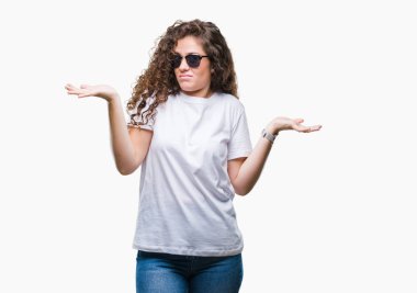Güzel esmer kıvırcık saçlı Genç kız üzerinde güneş gözlüğü takıyor arka plan clueless ve şaşkın bir ifadeyle kollar ve eller kaldırdı izole. Şüphe kavramı.