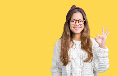 Gözlük ve kış şapka gülümseyen izole arka plan üzerinde el ve parmaklar ok işaretiyle yapıyor olumlu giyen genç güzel esmer hipster kadın. Başarılı ifade.