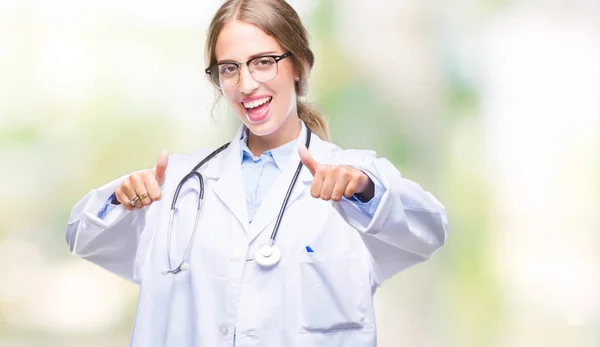 Beautiful young blonde doctor woman wearing medical uniform over isolated background approving doing positive gesture with hand, thumbs up smiling and happy for success. Looking at the camera, winner gesture.