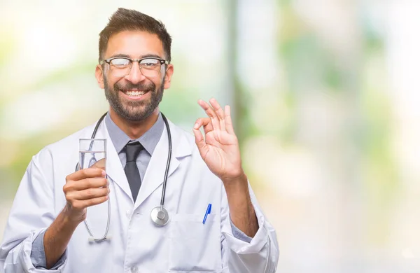 優秀なシンボルの指で サインをして孤立した背景に水のガラスを飲む大人のヒスパニック系医師男 — ストック写真