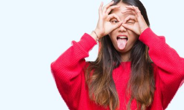 Genç Asyalı kadın dilini, parmaklarının arasından baktığını yapışmasını dürbün gibi ok jest yapıyor izole arka plan üzerinde kış kazak giyiyor. Deli ifade.