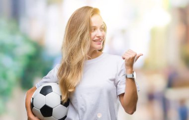 İşaret ve başparmak kadar mutlu yüz gülümseyen tarafı ile gösteren izole arka plan üzerinde futbol topu tutan genç güzel sarışın kadın
