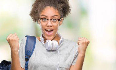 Kulaklıklar ve sırt çantası izole arka plan kutlama üzerinde giyen genç afro Amerikan öğrenci kadın şaşırttı ve başarı için kaldırdı kollarını açarak hayret ve gözleri açık. Kazanan kavramı.