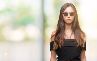 Genç Asyalı kadın yüzünde ciddi bir ifade ile izole arka plan üzerinde güneş gözlüğü takıyor. Basit ve doğal kameraya bakıyor.