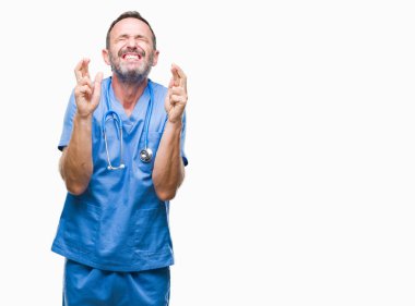 Geçiş parmakları umut ve gözleri kapalı ile gülümseyen izole arka plan üzerinde tıbbi üniforma giyen orta yaş ağarmış kıdemli Doktor adam. Şans ve batıl kavramı.