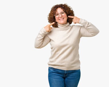 Boğazlı kazak ve gözlük emin gösterilen ve işaret parmakları diş ve ağız ile gülümseyen izole arka plan üzerinde giyen güzel orta ager üst düzey kadın. Sağlık kavramı.