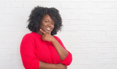Genç Afro-Amerikan artı boyutu kadın emin çapraz kollar ve çeneme kaldırdı el ile gülümseme ile kameraya bakarak beyaz tuğla duvarın üzerinden. Pozitif düşünce.