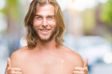 Genç yakışıklı gömleksiz adam uzun saçlı seksi vücut izole arka plan başarı işareti el ile pozitif jest yapıyor üzerinde gösterilen başparmak gülümseyen ve mutlu. Neşeli bir ifadeyle, kazanan jest kameraya bakıyor.