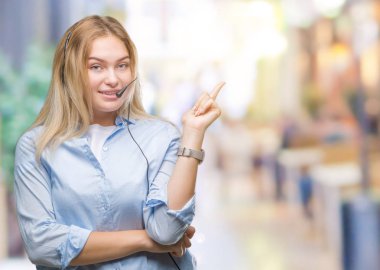 Yüzünde kocaman bir gülümseme ile izole arka plan üzerinde çağrı merkezi kulaklık giyen genç beyaz iş kadın kameraya bakarak yan el ve parmak ile işaret.