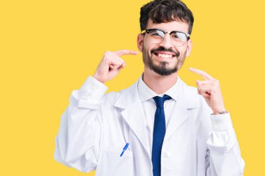 Kendinden emin gösterilen ve işaret parmakları diş ve ağız ile gülümseyen izole arka plan üzerinde beyaz önlük giyen genç profesyonel bilim adam. Sağlık kavramı.