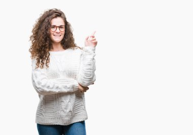 Kameraya bakarak yan el ve parmak ile işaret yüzünde kocaman bir gülümseme ile izole arka plan üzerinde kış kazak giyen güzel esmer kıvırcık saçlı Genç kız.