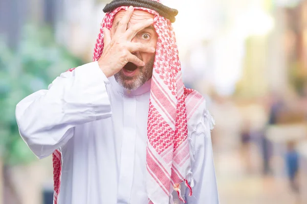 恥ずかしそうな顔で指を通して見る顔と手で目を覆うショックでピーク分離の背景にクーフィーヤを着てシニアのアラブ人 — ストック写真