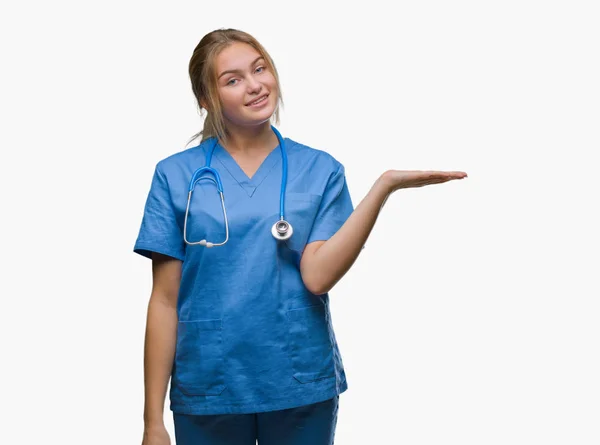 年轻的白种人医生妇女穿外科医生制服在孤立的背景微笑欢快的呈现和指向手掌的手看着相机 — 图库照片