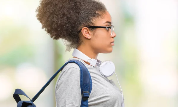 Young afro american student woman wearing headphones and backpack over isolated background looking to side, relax profile pose with natural face with confident smile.