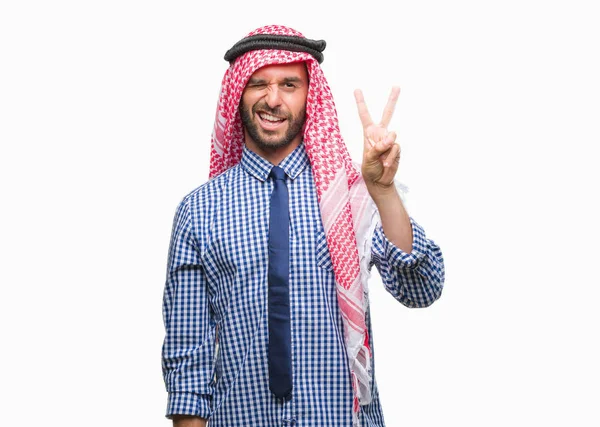 勝利のサインを行うカメラでウィンクしている幸せそうな顔を浮かべて隔離された背景にクーフィーヤを着た若いハンサムなアラビアン ビジネス男 — ストック写真
