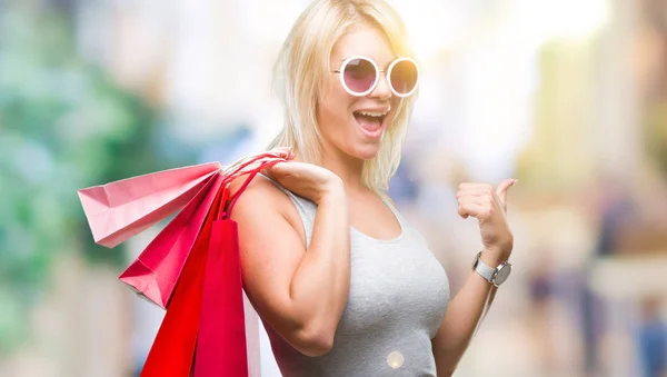 若いの美しいブロンドの女性を指していると幸せそうな顔を笑顔で側まで親指で示す分離の背景の上販売開催ショッピング バッグをショッピング — ストック写真