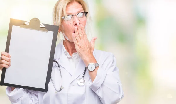 Middle age blonde doctor woman holding clipboard over isolated background cover mouth with hand shocked with shame for mistake, expression of fear, scared in silence, secret concept