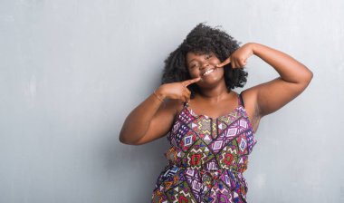 Afrikalı-Amerikalı genç kadın kendine güvenen gösterilen ve işaret parmakları diş ve ağız ile gülümseyen renkli elbise gri grunge duvarın üzerinden. Sağlık kavramı.