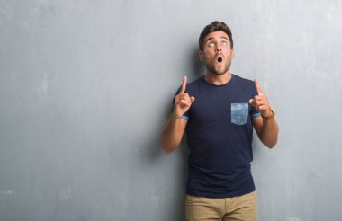 Yakışıklı genç gri grunge üzerinden hayran ve sürpriz ve parmakları ile işaret duvar ve silah kaldırdı.