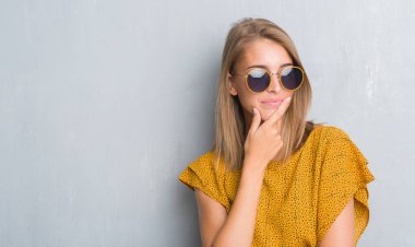 Retro güneş gözlüğü ciddi bir yüz düşünme hakkında soru, çok karışık fikir giyen grunge gri duvar üzerinde duran güzel genç kadın