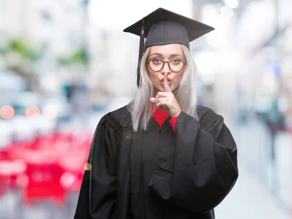 若いブロンドの女性は 唇に指で静かにするように求めて孤立の背景の上の大学院の制服を着ています 沈黙と秘密の概念 — ストック写真