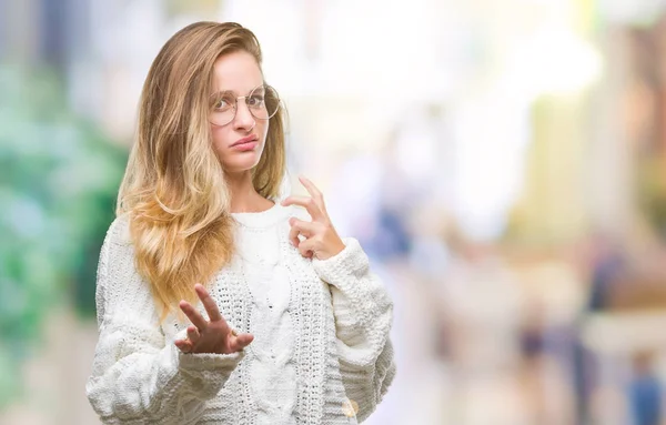 Young beautiful blonde woman wearing winter sweater and sunglasses over isolated background disgusted expression, displeased and fearful doing disgust face because aversion reaction. With hands raised. Annoying concept.