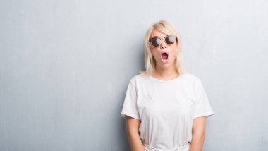 Grunge gri duvar korkuyor ve şok sürpriz ifade, korku ve heyecan yüz ile güneş gözlüğü takıyor yetişkin beyaz kadın.