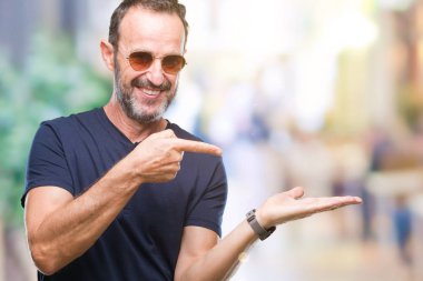 Orta yaş ağarmış komuta sizde hayrete ve el ile sunulması ve parmak ile işaret ederken kameraya gülümseyen izole arka plan üzerinde güneş gözlüğü takıyor.