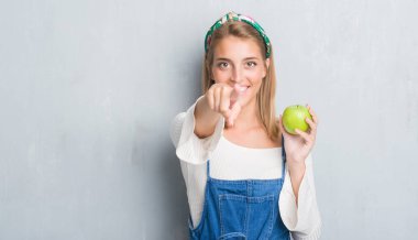 Güzel genç kadın kamera ve size parmak ile işaret yeşil elma yeme grunge gri duvarın üzerinden, el işareti, pozitif ve kendinden emin bir jest ön