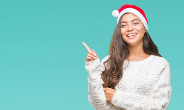 Kameraya bakarak yan el ve parmak ile işaret yüzünde kocaman bir gülümseme ile izole arka plan üzerinde Noel şapka giyen genç Arap kadın.