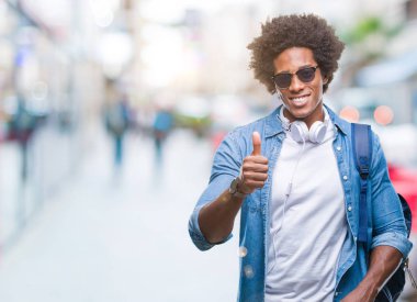 Afro Amerikalı adam ok işareti, başparmak ile parmak, mükemmel işareti yapıyor kocaman bir gülümseme ile izole arka plan üzerinde mutlu giyen kulaklık ve sırt çantası