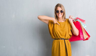Grunge gri duvar kendisine sürpriz yüz işaret parmağı satış alışveriş torbaları tutan güzel genç kadın