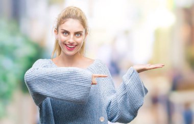 Hayret ve el ile sunulması ve parmak ile işaret ederken kameraya gülümseyen izole arka plan üzerinde kış kazak giyen genç güzel sarışın ve mavi gözlü kadın.