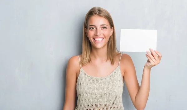 Beautiful young woman over grunge grey wall holding blank card with a happy face standing and smiling with a confident smile showing teeth