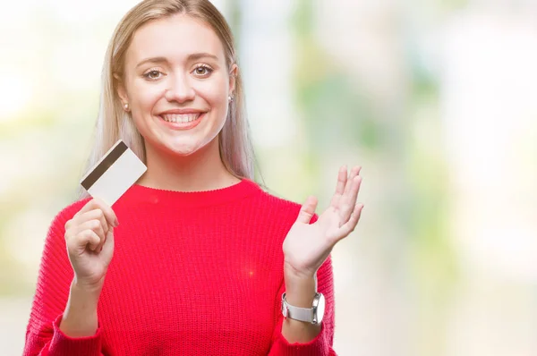 Young blonde woman holding credit card over isolated background very happy and excited, winner expression celebrating victory screaming with big smile and raised hands