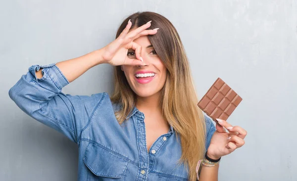 指の間から見て目に手で サインをして笑っている幸せそうな顔でチョコレートバーを灰色のグランジの壁を食べる以上の若い成人女性 — ストック写真